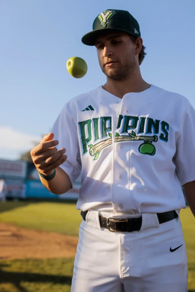 A Yakima Valley Pippins baseball player in a white jersey and green cap tossing a green apple into the air